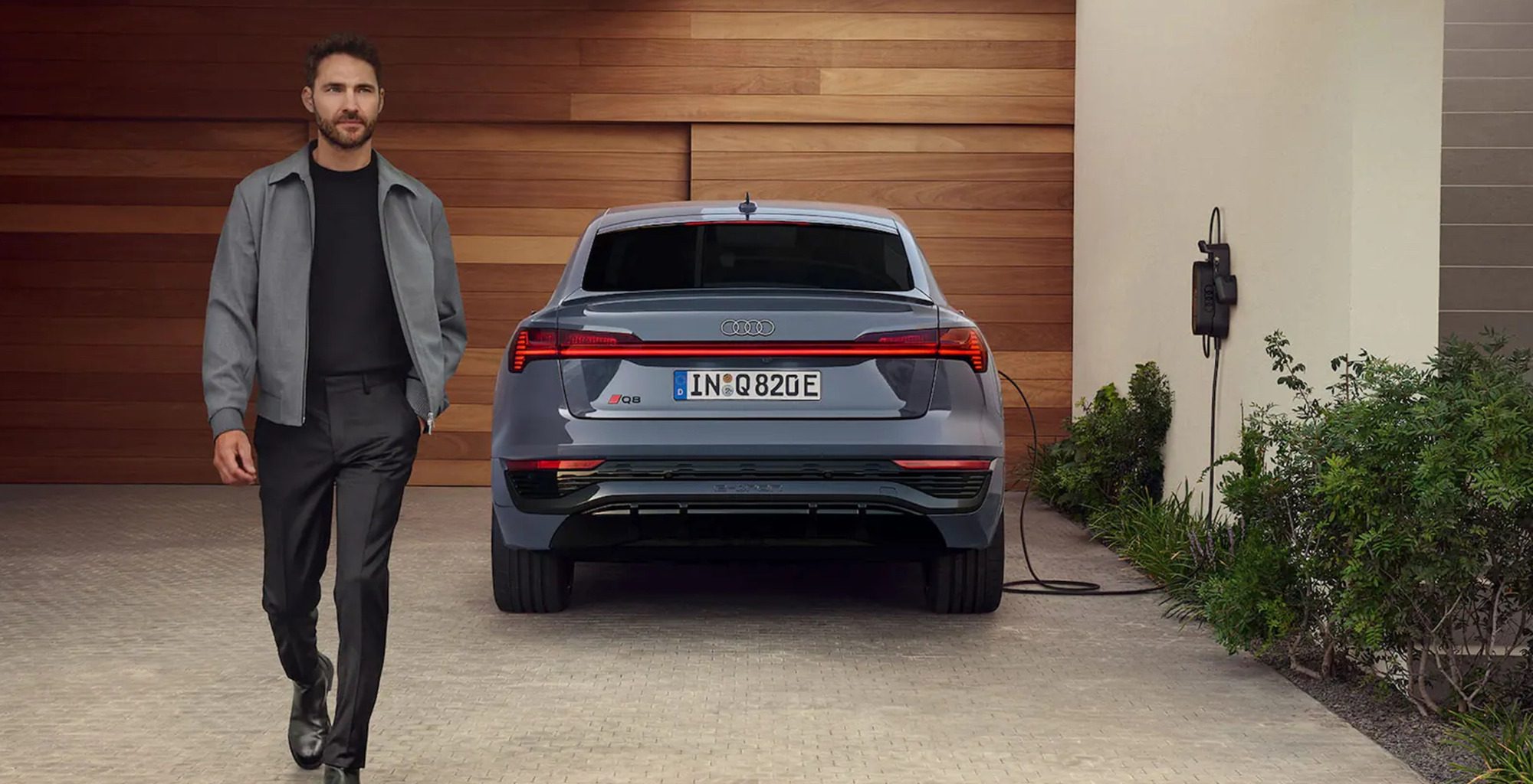 Grey Audi Sq8 e-tron parked in drive way plugged in to a charger with a man in casual clothes to the left